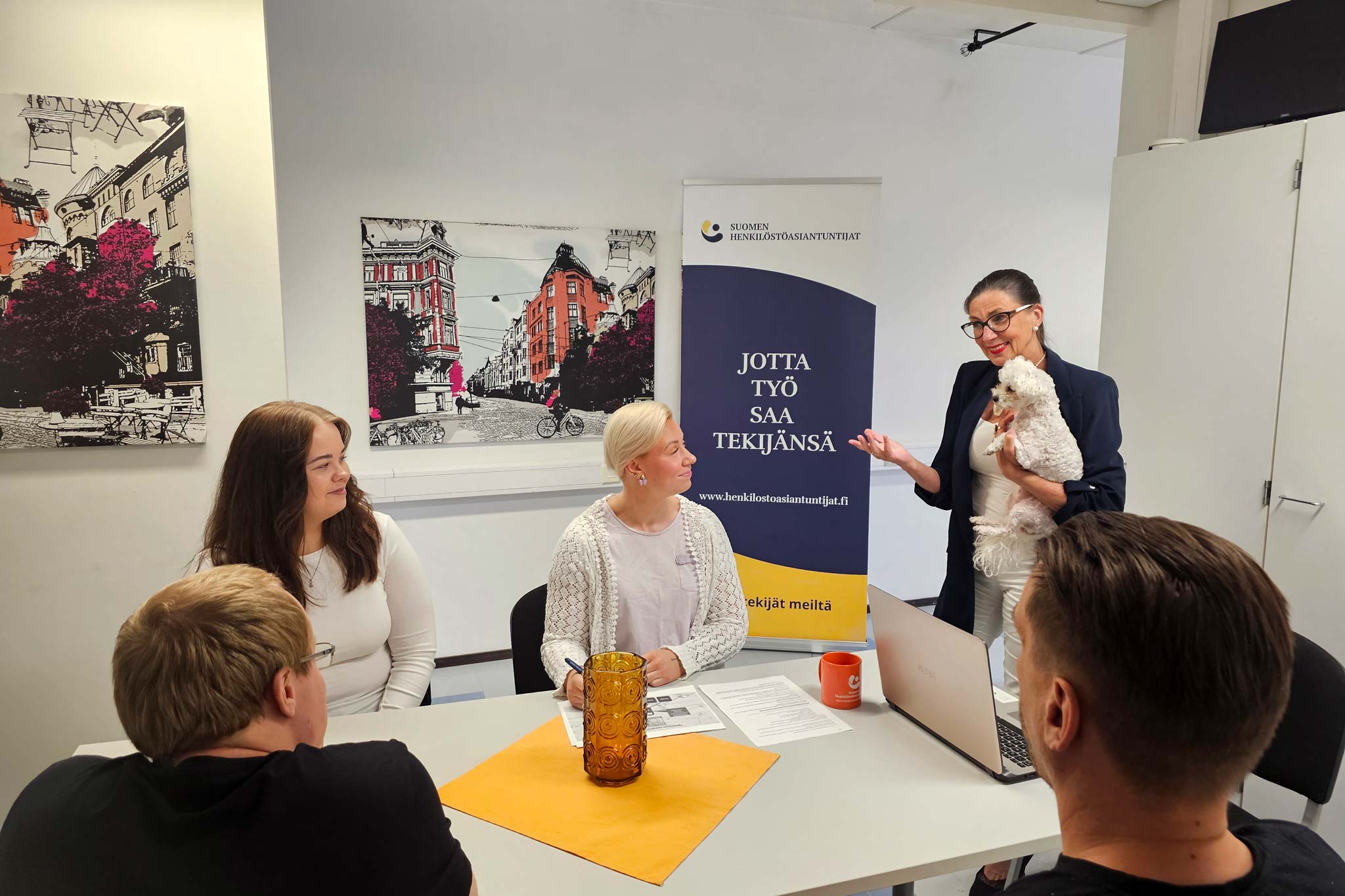 Rekrytointitilanteessa henkilöstöasiantuntija esittelee palvelua pienelle ryhmälle toimistossa, sylissään pieni koira.